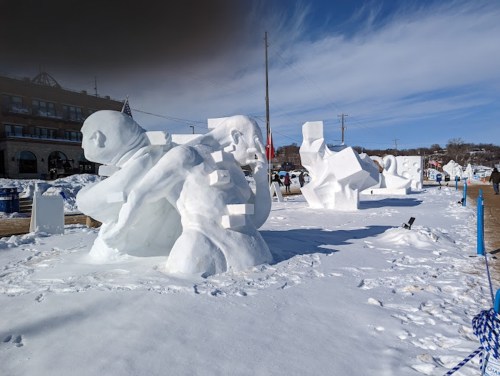 Snow sculptures of various abstract shapes and figures in a snowy landscape, with buildings in the background.