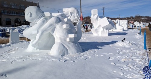 Snow sculptures in a snowy landscape, featuring abstract and human-like forms against a clear blue sky.