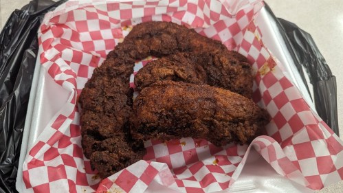 Three pieces of crispy fried chicken on a red and white checkered paper in a takeout container.