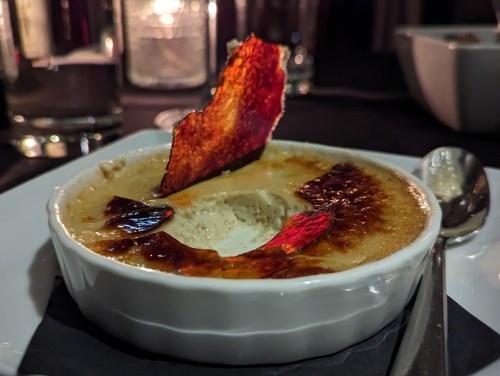 A creamy dessert in a white dish, topped with caramelized sugar and a decorative shard, set on a dark table.