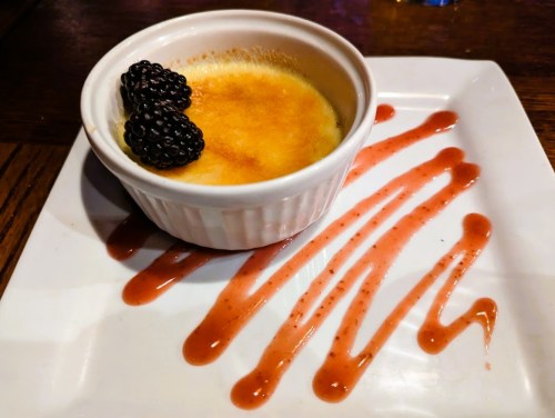A creamy dessert in a ramekin topped with blackberries, served on a plate with raspberry sauce drizzled around it.