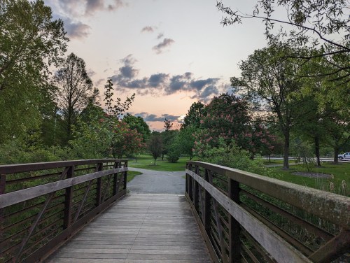 A wooden bridge leads into a park at sunset, surrounded by trees and colorful clouds in the sky.