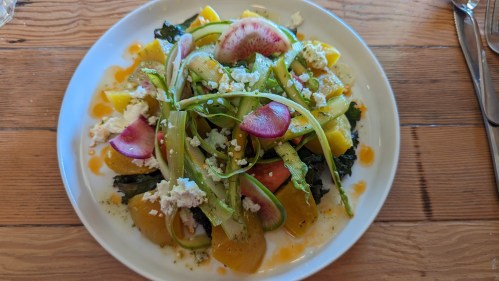A colorful salad with greens, yellow beets, radishes, and feta cheese, drizzled with a light dressing on a white plate.
