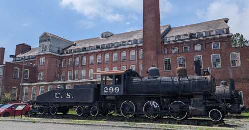 A vintage steam locomotive labeled "U.S. 299" parked beside a large brick building under a blue sky.