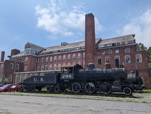 An old steam locomotive labeled "U.S. 299" parked beside a large, abandoned brick building under a blue sky.