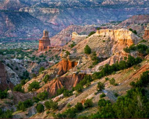 A scenic view of colorful canyons and rock formations under a clear sky, with greenery scattered throughout the landscape.