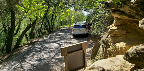 A parked car on a narrow road surrounded by trees and rocky terrain, with a wooden sign in the foreground.