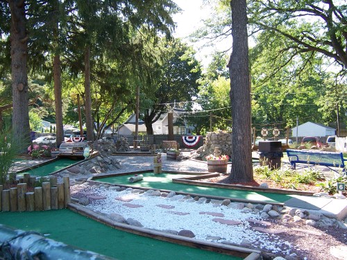A scenic mini-golf course with green turf, stone features, and trees, set in a sunny outdoor environment.