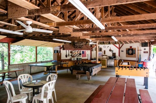 Interior of a rustic lodge with wooden beams, pool table, foosball table, and picnic-style seating. Natural light filters in.