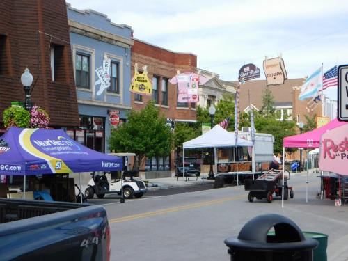 A street scene featuring colorful storefronts, food trucks, and tents, with banners and flags decorating the area.