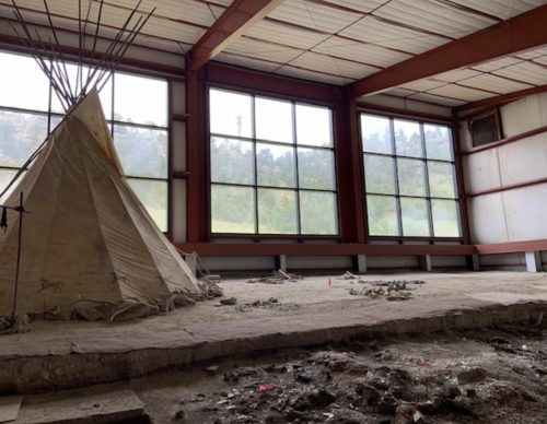 Interior of a large, unfinished space featuring a teepee and large windows with a view of greenery outside.