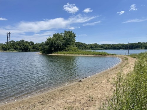 A serene lakeside view with sandy shore, lush greenery, and a clear blue sky dotted with clouds.