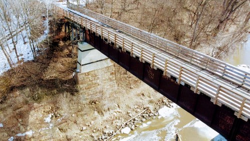 Aerial view of a wooden bridge over a river, surrounded by trees and snow, with rocky banks visible below.