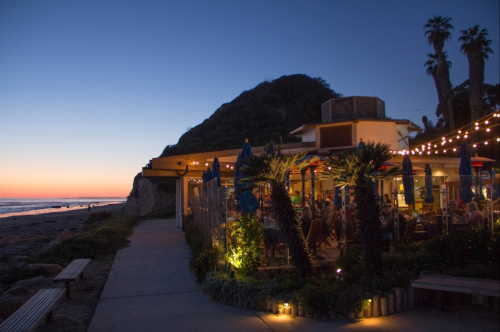 Cozy beachside restaurant at dusk, with string lights and palm trees, overlooking the ocean and a sunset.