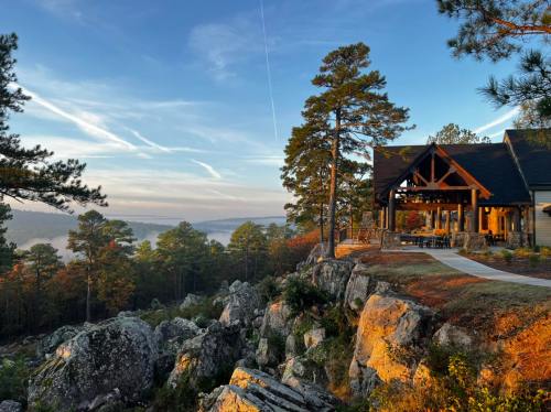 A scenic view of a cabin on a rocky hillside, surrounded by trees and a misty valley at sunrise.