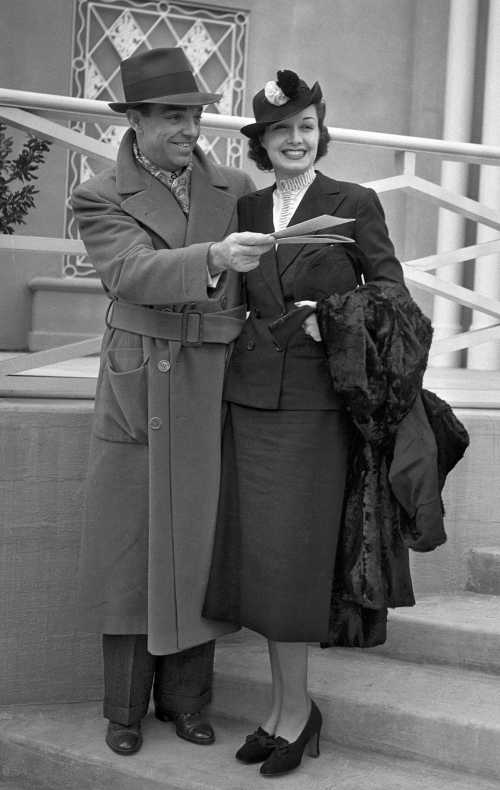 A man in a coat and hat stands beside a woman in a stylish outfit, both smiling and holding a piece of paper.