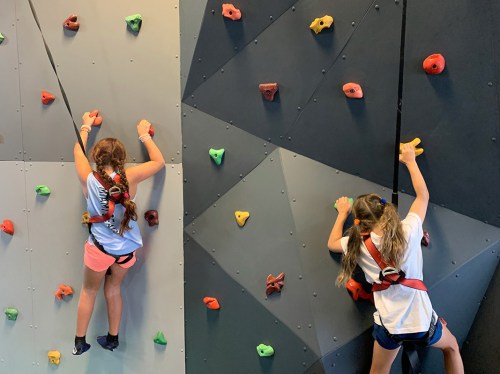Two children climbing a colorful indoor rock wall, wearing harnesses and focused on reaching the top.