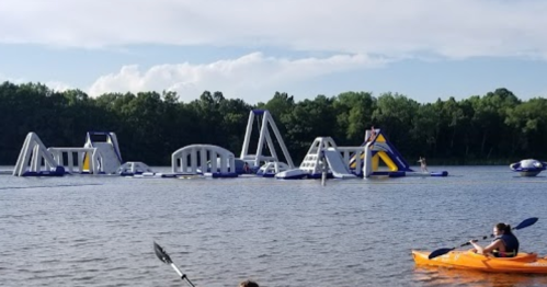 A person in a kayak paddles near a large inflatable water park on a lake, surrounded by trees and blue skies.