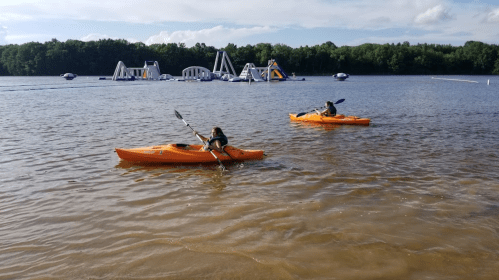 Two people kayak on a calm lake with inflatable water structures in the background under a clear blue sky.