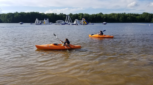 Two people kayaking on a lake with inflatable water structures in the background under a clear blue sky.