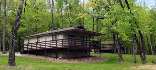 A single-story house with a wooden deck, surrounded by lush green trees in a wooded area.
