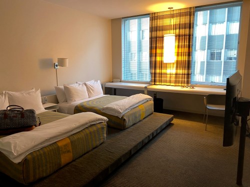 A modern hotel room with two beds, a large window, a desk, and a TV, featuring minimalist decor and warm lighting.