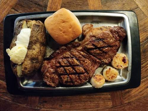 A grilled steak dinner featuring two steaks, a baked potato, a roll, and seared scallops on a metal tray.