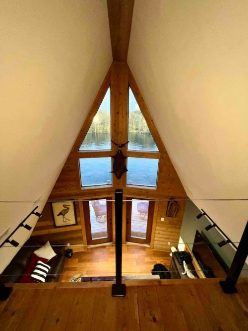 A cozy wooden cabin interior with a triangular window overlooking a lake, featuring a modern design and warm lighting.