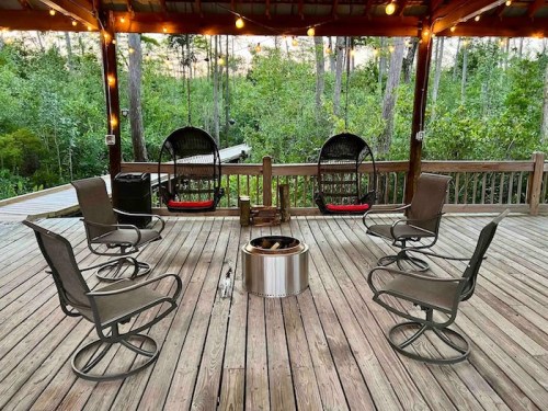 A cozy outdoor seating area with six chairs around a fire pit, surrounded by trees and string lights.