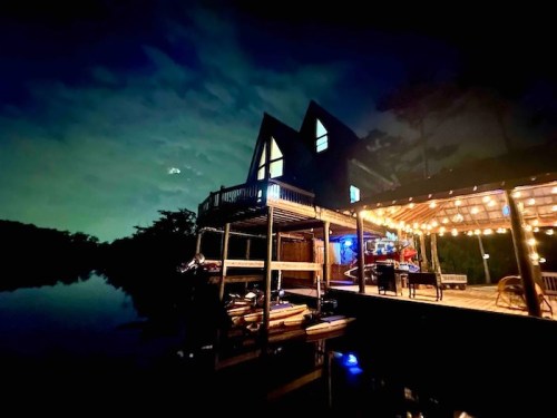 A cozy house by the water at night, illuminated by string lights under a starry sky.