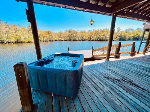 A hot tub on a wooden deck overlooking a calm river surrounded by trees under a clear blue sky.