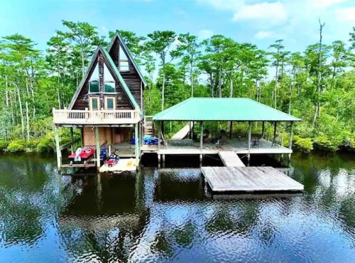 Aerial view of a triangular cabin by a calm waterway, surrounded by lush greenery and a wooden dock.