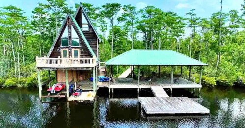 Aerial view of a triangular cabin by a calm river, surrounded by lush green trees and a wooden dock.