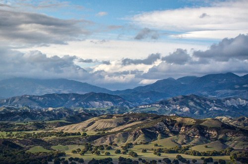 A scenic view of rolling hills and mountains under a cloudy sky, showcasing lush greenery and varied terrain.