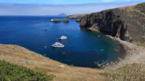 A serene coastal view with boats anchored in a calm bay, surrounded by rocky cliffs and grassy hills under a clear sky.