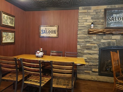 A rustic dining area with a wooden table, chairs, and a stone fireplace, featuring saloon-themed decor on the walls.