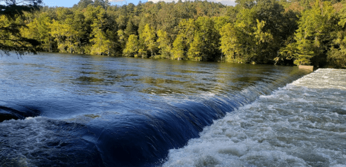 A serene river scene with a gentle waterfall cascading over a dam, surrounded by lush green trees in autumn colors.