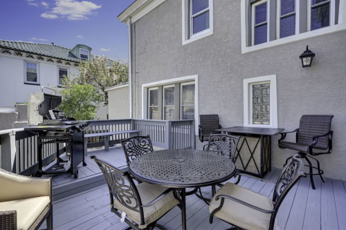 A spacious outdoor deck with a dining table, chairs, and a grill, surrounded by buildings and trees.