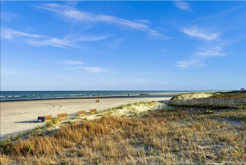 A serene beach scene with gentle waves, sandy dunes, and a clear blue sky. A few beach chairs are visible in the distance.