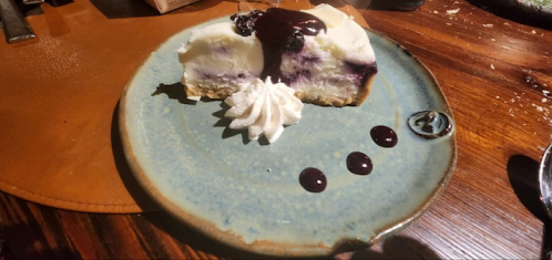 A slice of cheesecake with blueberry topping, whipped cream, and chocolate drizzle on a rustic plate.