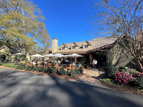 A charming restaurant with outdoor seating, surrounded by trees and flowers, under a clear blue sky.