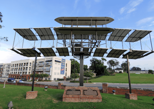 A large metal sculpture of an early airplane on display in a grassy area near a building.