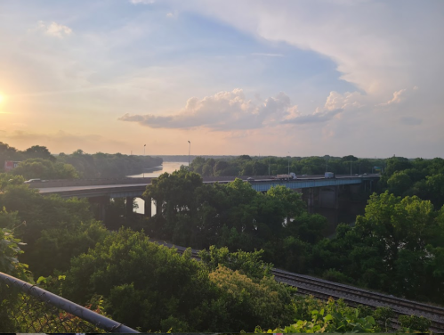 A scenic view of a river and bridge at sunset, surrounded by lush greenery and a cloudy sky.