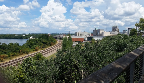 A scenic view of a river, railway tracks, and a city skyline under a partly cloudy sky. Lush greenery in the foreground.