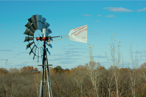 A vintage windmill stands against a blue sky, surrounded by trees with autumn foliage.