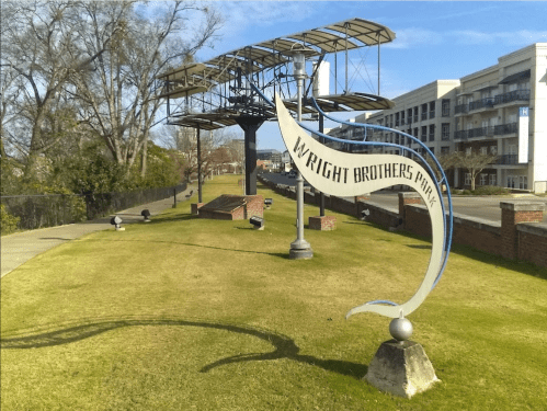 Sign for Wright Brothers Park with a historic airplane structure in the background and green grass in the foreground.