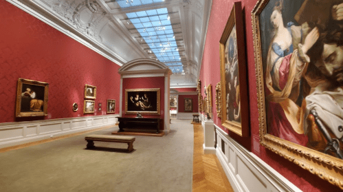 A spacious art gallery with red walls, featuring classic paintings and a wooden bench in the center.