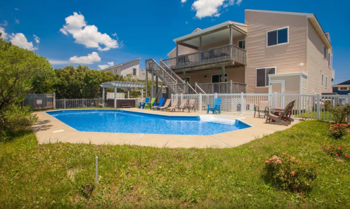 A sunny backyard with a swimming pool, surrounded by green grass and a house with a deck and seating area.
