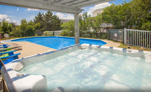 A hot tub in the foreground with a blue swimming pool and greenery in the background under a clear blue sky.