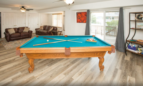 A spacious room featuring a blue pool table, two couches, and large windows with a view of a deck outside.